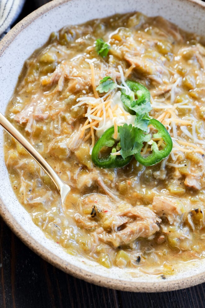 A bowl of pork green chile stew with shredded meat, garnished with sliced jalapeños, shredded cheese, and cilantro, with a spoon resting inside.