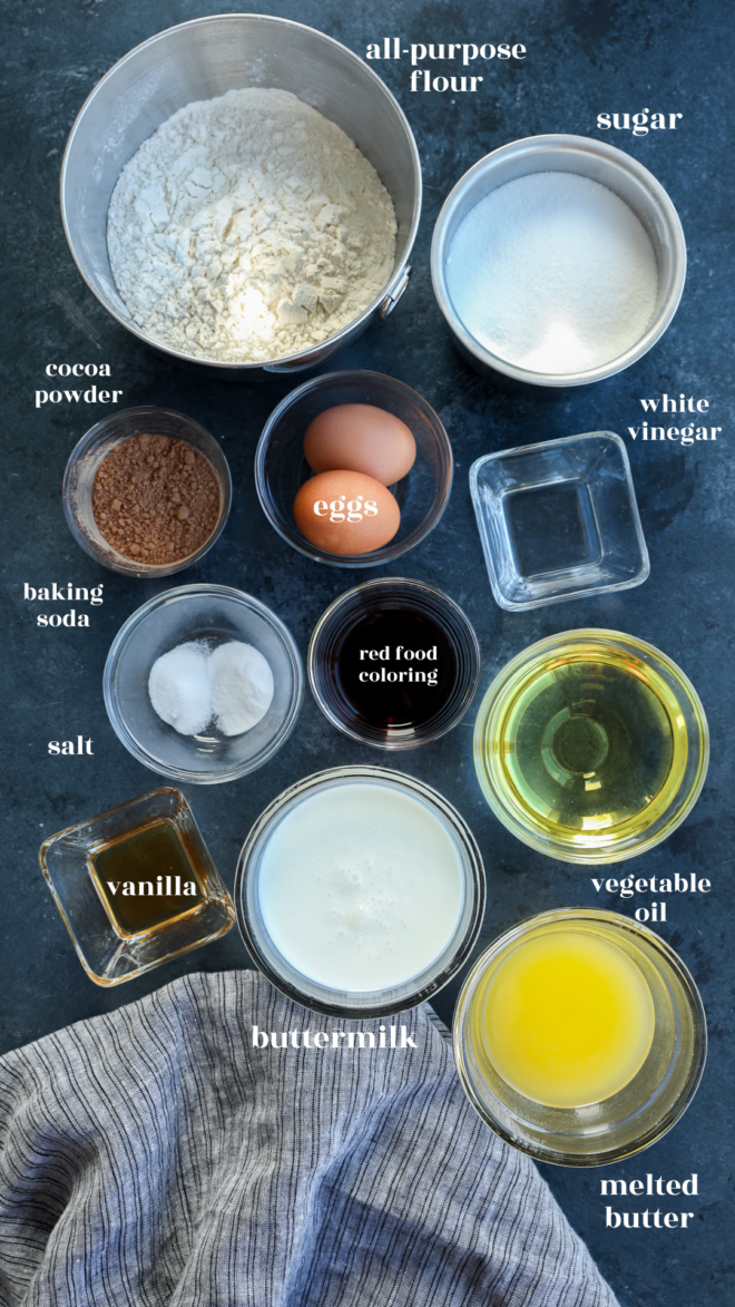 Overhead view of labeled baking ingredients in bowls and cups for Red Velvet Bundt Cake, including flour, sugar, cocoa powder, baking soda, salt, vanilla, eggs, buttermilk, red food coloring, white vinegar, vegetable oil, and melted butter.