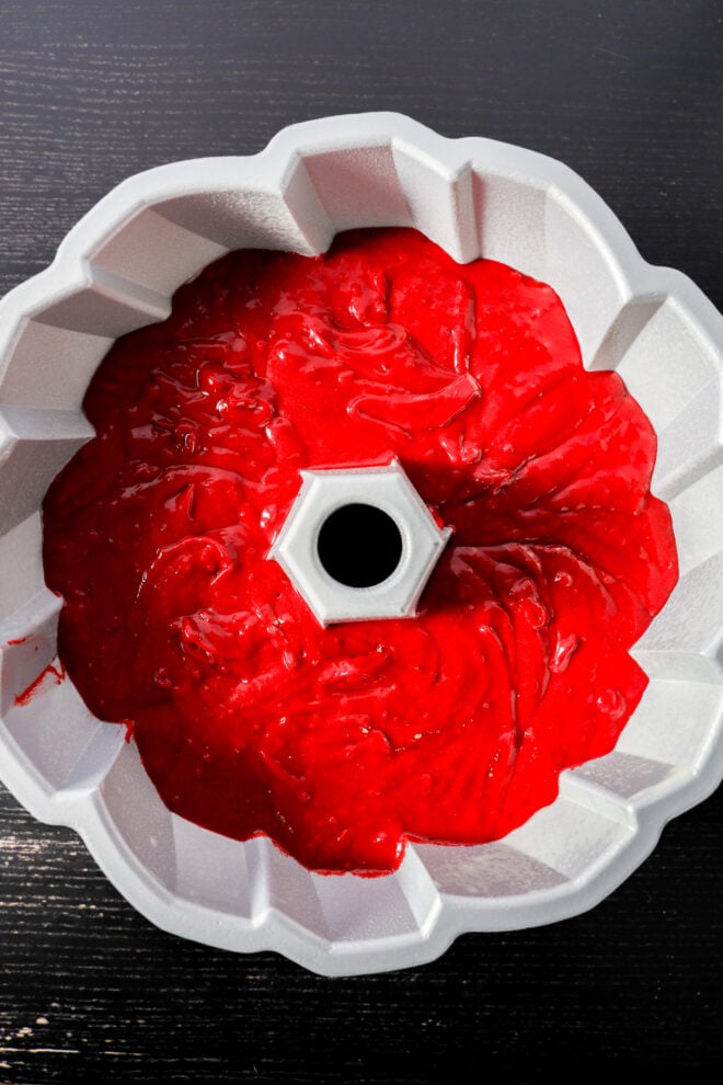 A Red Velvet Bundt Cake pan filled with bright red cake batter sits on a dark wooden surface, ready to be baked.