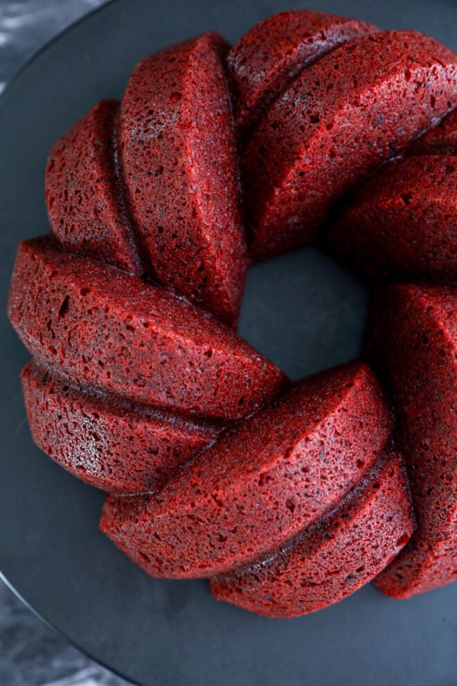 A close-up of a Red Velvet Bundt Cake with a swirl pattern, displayed on a dark plate. The cake has a rich red color and a moist, textured surface.
