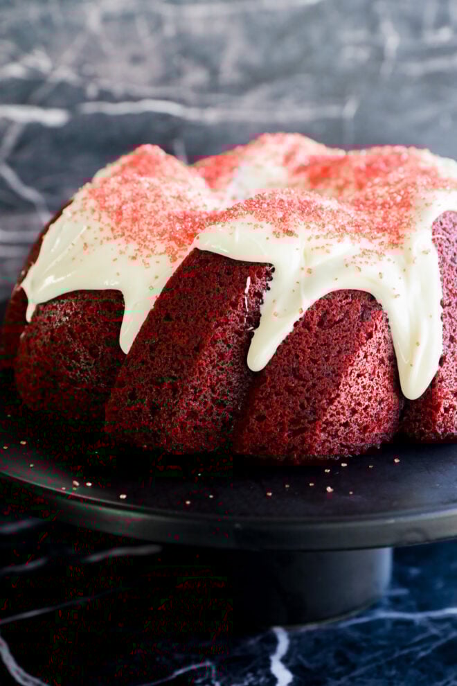 A classic Red Velvet Bundt Cake with white icing drizzled on top and red sprinkles, beautifully displayed on a black cake stand against a dark marble background.