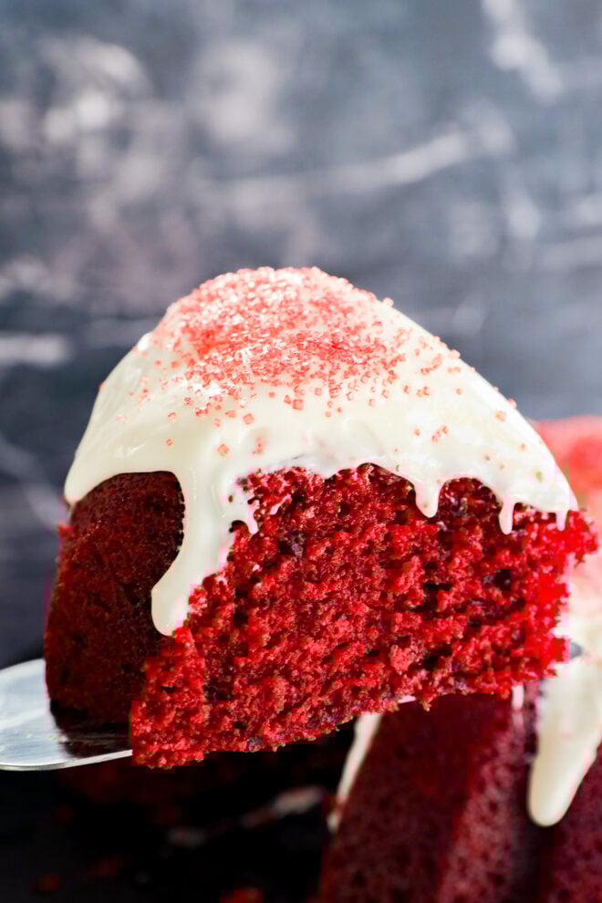 A close-up of a Red Velvet Bundt Cake slice with white icing drizzling down the sides and red sugar sprinkles on top, set against a dark, blurred background.