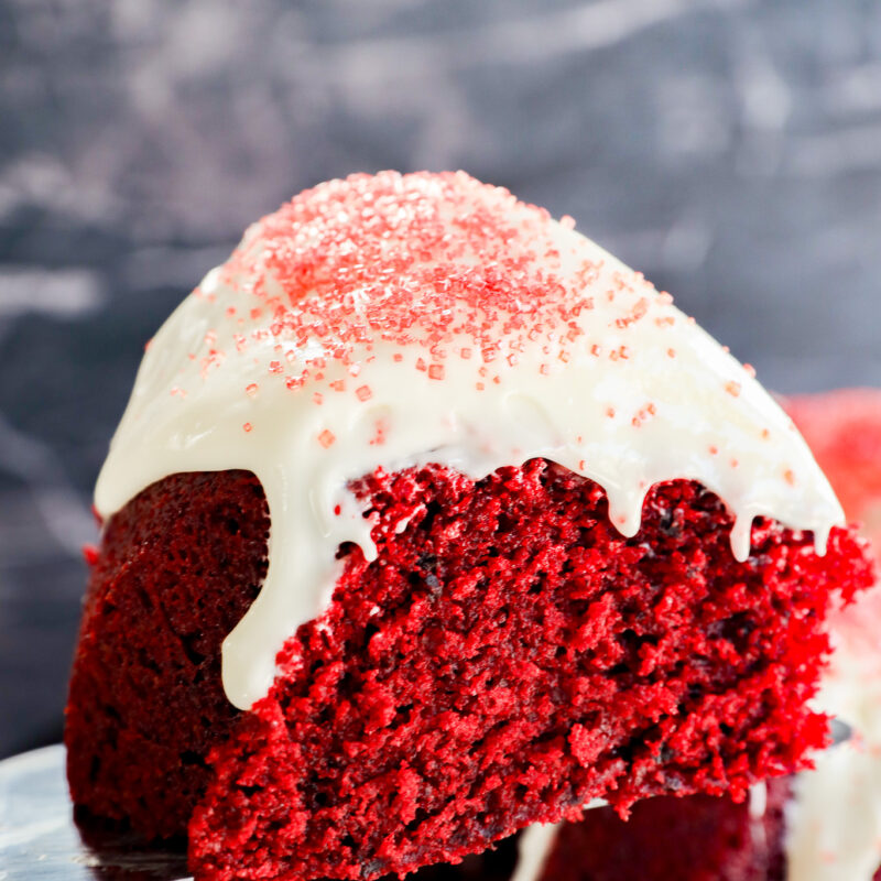 A close-up of a Red Velvet Bundt Cake slice with white icing drizzling down the sides and red sugar sprinkles on top, set against a dark, blurred background.