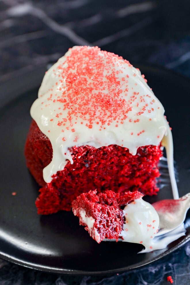 A slice of Red Velvet Bundt Cake topped with white icing and red sprinkles sits on a black plate, with a fork holding a bite-sized piece in the foreground.