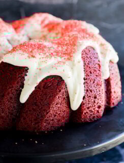 A close-up of a Red Velvet Bundt Cake topped with white icing and red sprinkles, elegantly displayed on a dark serving plate against a rich, dark background.