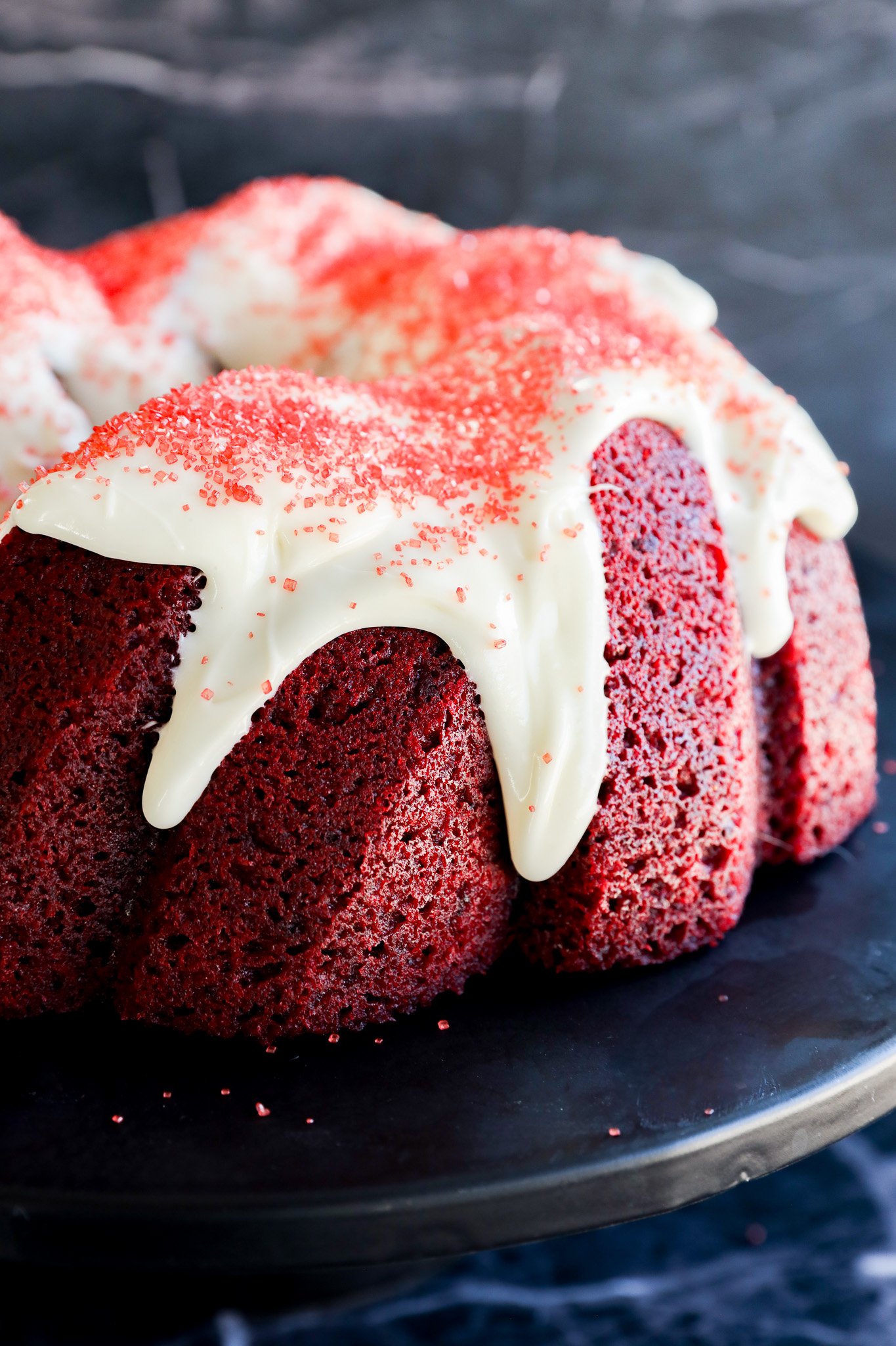 A close-up of a Red Velvet Bundt Cake topped with white icing and red sprinkles, elegantly displayed on a dark serving plate against a rich, dark background.