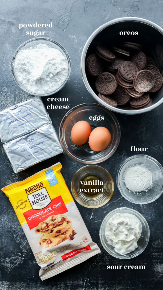 An overhead view of ingredients for slutty cheesecake bars in bowls and packages&mdash;powdered sugar, Oreos, cream cheese, eggs, flour, vanilla extract, chocolate chip morsels, and sour cream&mdash;all arranged on a dark surface.