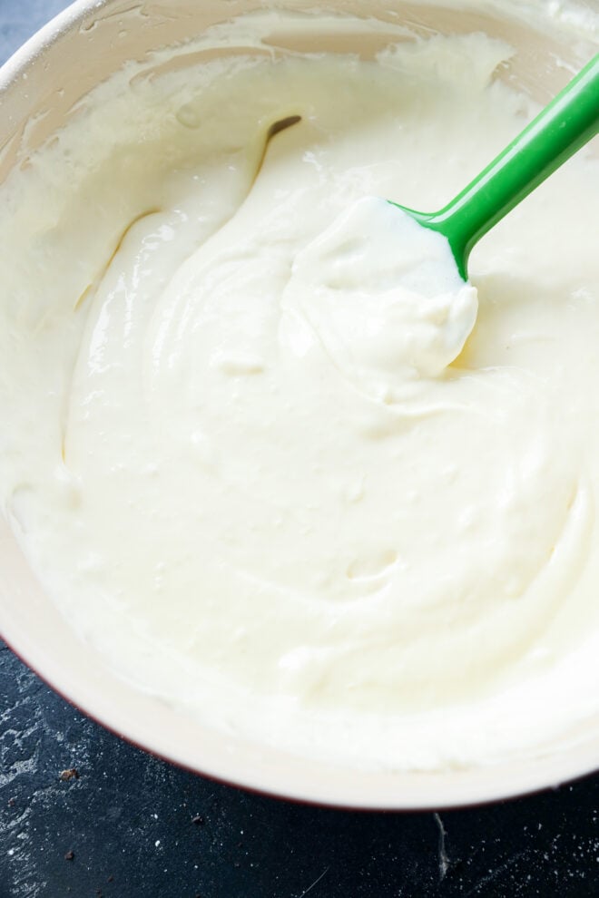 A green spatula is stirring thick, creamy white batter in a large mixing bowl.