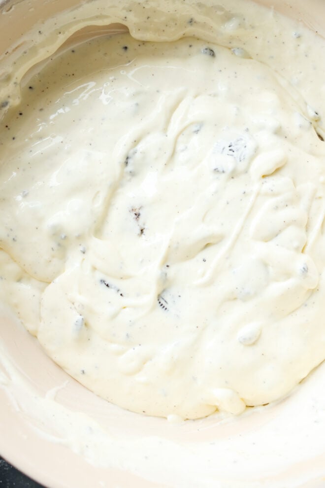Close-up of a creamy white mixture with visible specks and small chunks, likely the rich batter with crushed cookies or chocolate pieces stirred in.