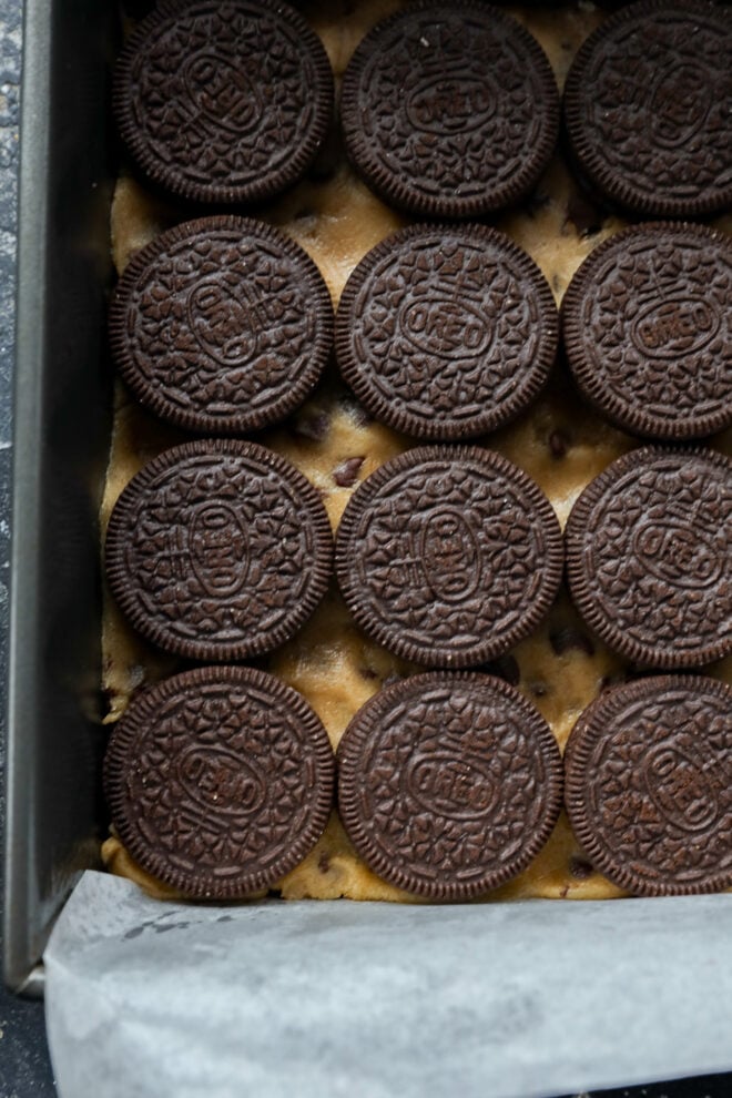 A baking pan lined with parchment paper contains a layer of chocolate chip cookie dough topped with rows of chocolate sandwich cookies