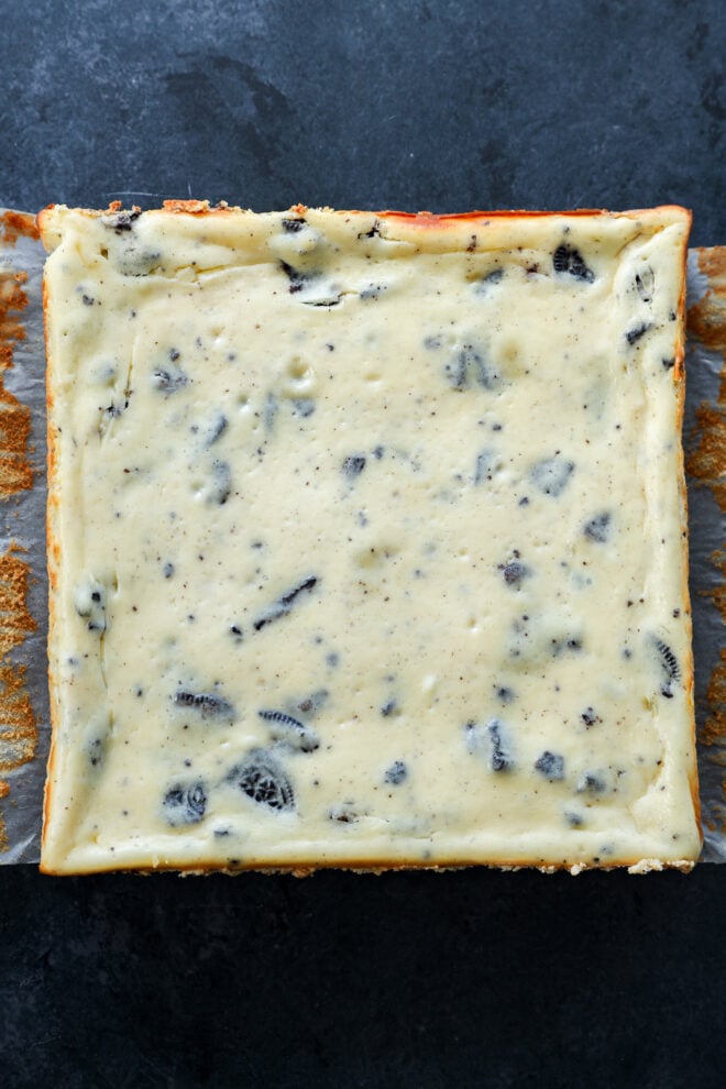 A square pan of slutty cheesecake bars with a creamy cookies and cream filling and crushed cookie pieces, set on a dark surface with parchment paper lining the pan.