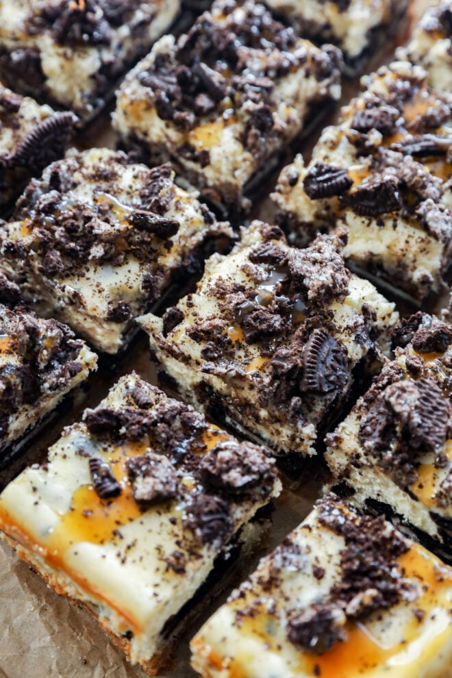 Close-up of slutty cheesecake bars topped with crushed chocolate cookies and drizzled caramel, cut into squares and arranged in neat rows on a sheet of parchment paper.