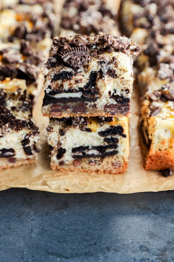 Two stacked slutty cheesecake bars with layers of cookie dough, cheesecake, chocolate chips, and crumbled chocolate sandwich cookies rest on parchment paper, with more decadent bars in the background.