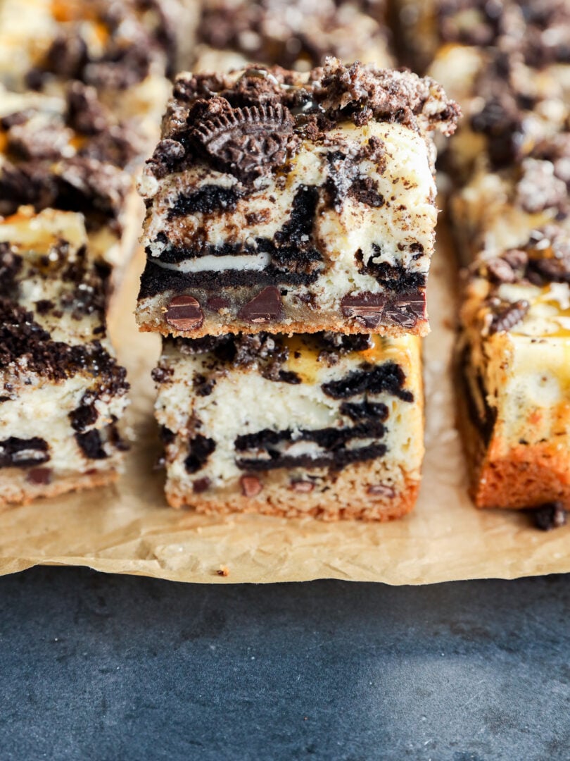 Two stacked slutty cheesecake bars with layers of cookie dough, cheesecake, chocolate chips, and crumbled chocolate sandwich cookies rest on parchment paper, with more decadent bars in the background.