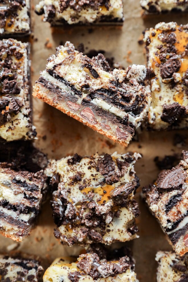 Close-up of slutty cheesecake bars topped with crushed chocolate cookies and drizzled with caramel, showing decadent, layered chocolate and cookie crusts on parchment paper.