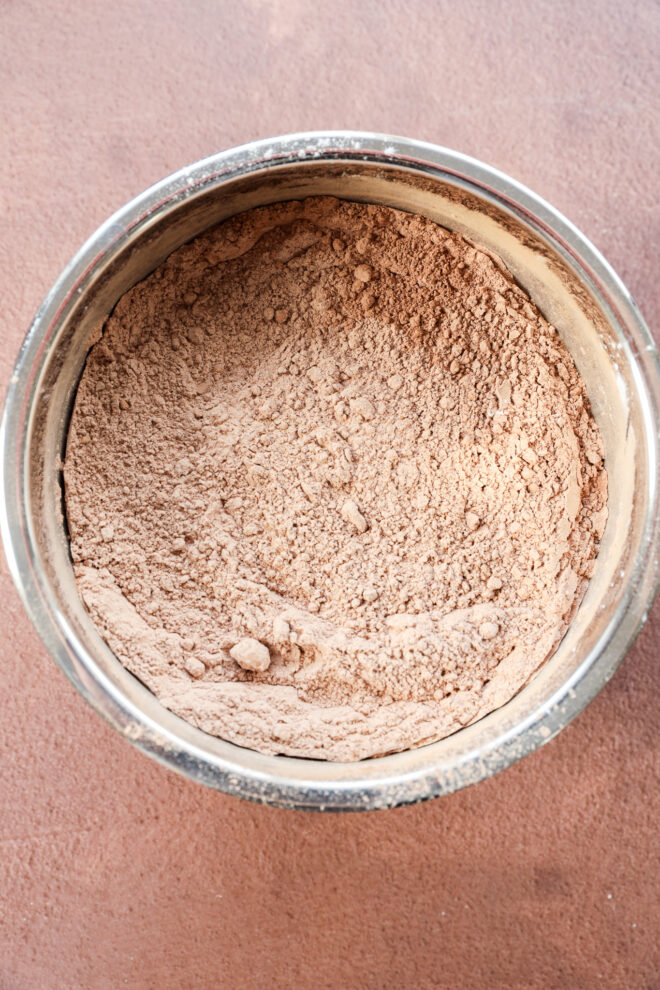A metal bowl filled with light brown cocoa powder, sits on a reddish-brown surface. The powder is finely textured with a few small clumps visible.