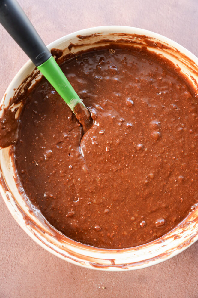 A bowl of brownie cake batter being mixed with a green-handled spatula on a light brown surface.