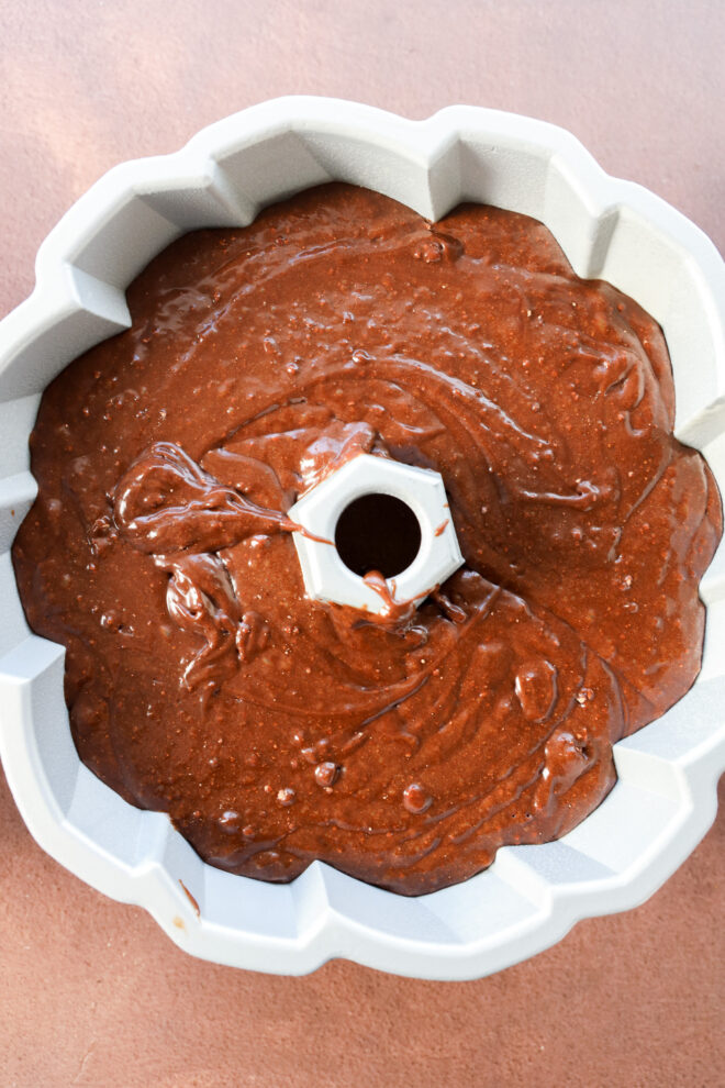 Brownie cake batter poured into a white Bundt pan, ready to be baked, on a light brown surface.