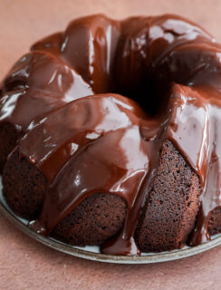 A rich brownie cake in bundt form, covered with glossy chocolate glaze, sits on a round metal plate against a brown background.