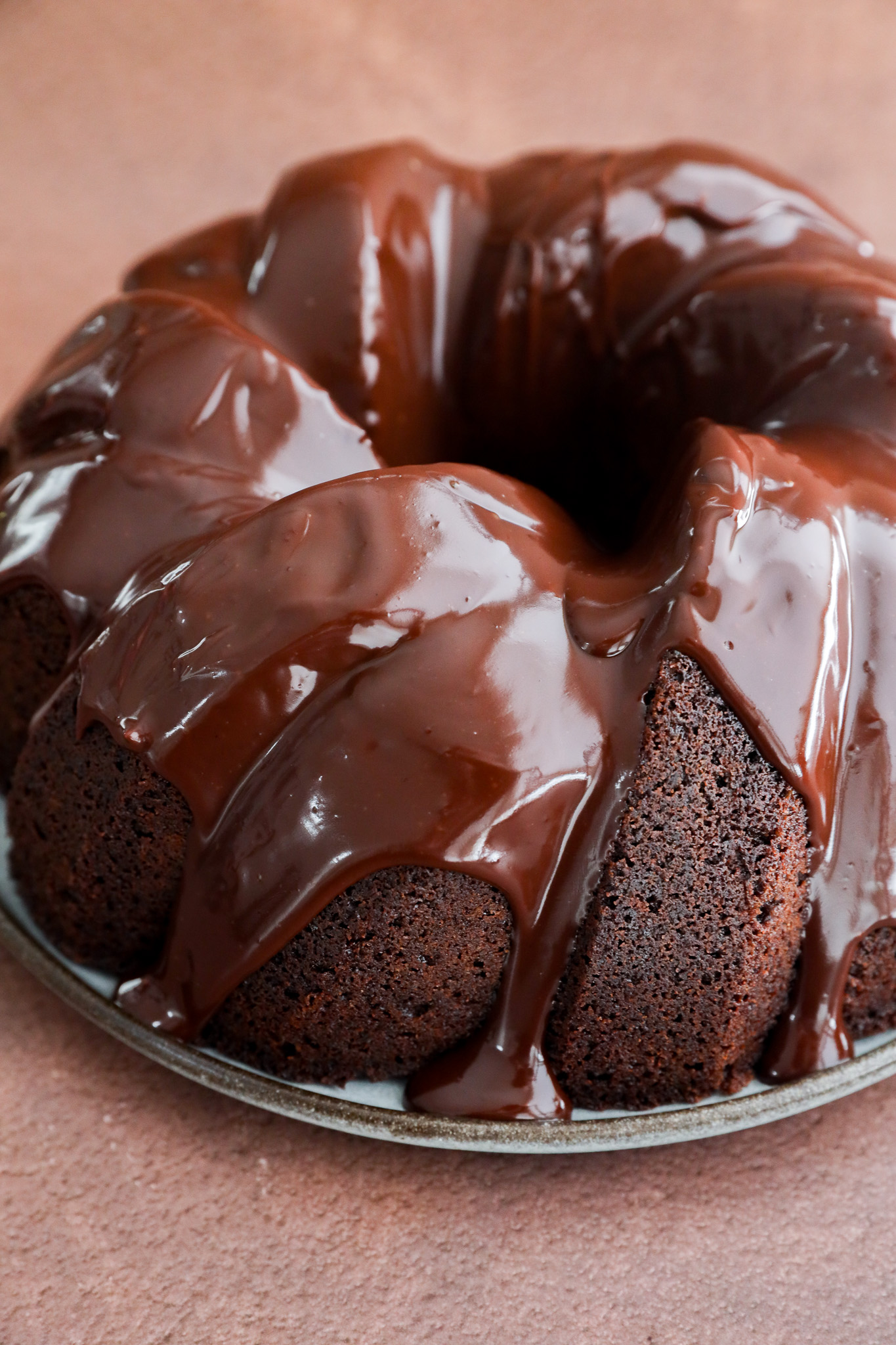 A rich brownie cake in bundt form, covered with glossy chocolate glaze, sits on a round metal plate against a brown background.