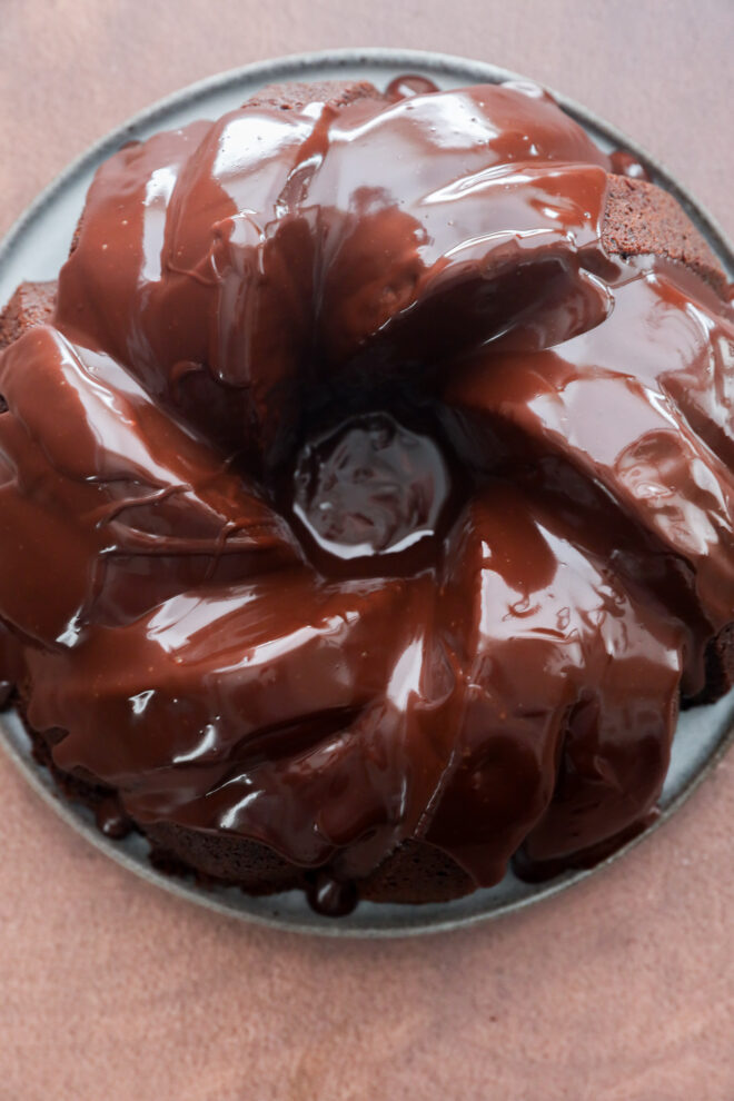 A brownie cake bundt covered in glossy, rich chocolate ganache sits on a round plate against a brown background.