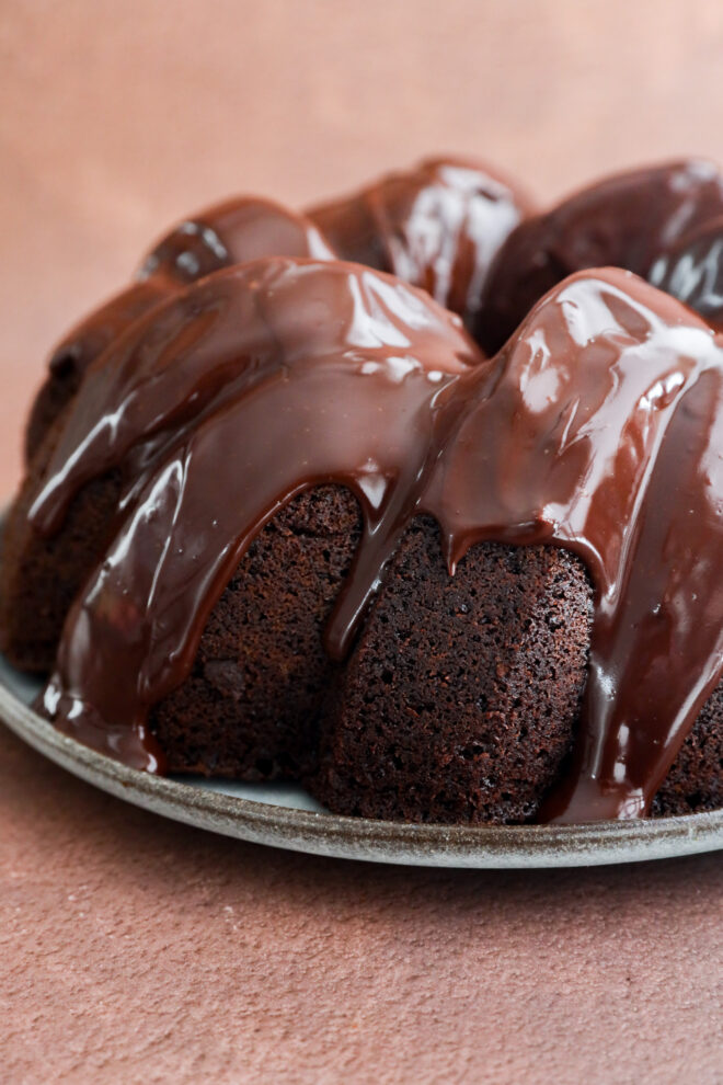 A close-up of a chocolate bundt cake, resembling a rich brownie cake, drizzled with glossy chocolate glaze and sitting on a plate against a light brown background.