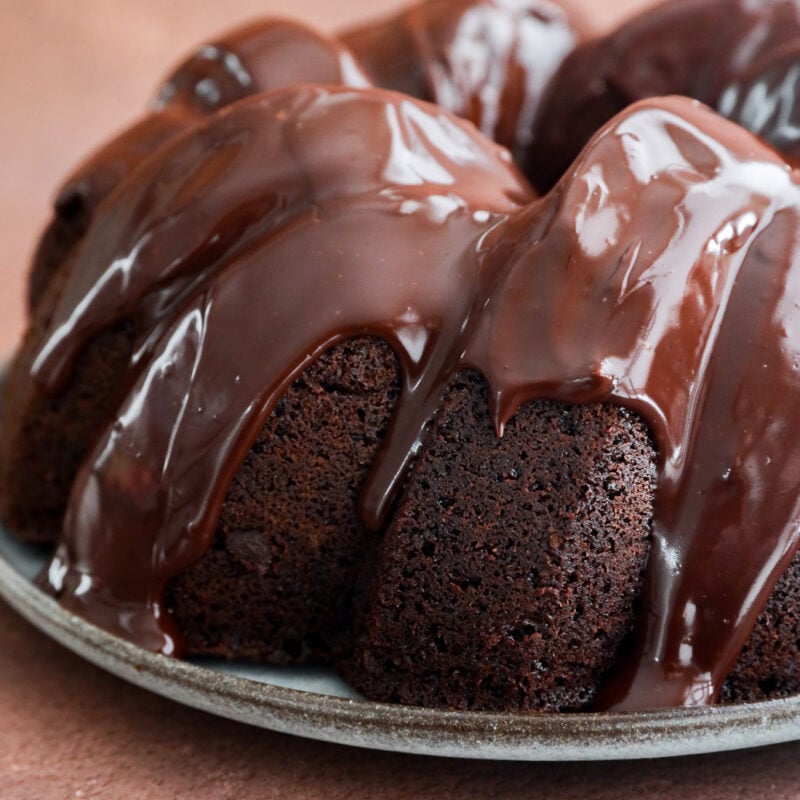 A close-up of a chocolate bundt cake, resembling a rich brownie cake, drizzled with glossy chocolate glaze and sitting on a plate against a light brown background.