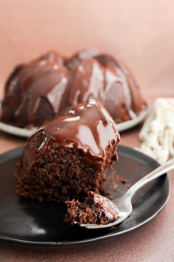 A close-up of a slice of chocolate bundt cake, reminiscent of a brownie cake, with glossy chocolate glaze on a black plate and a forkful in front. The remaining cake is blurred in the background.