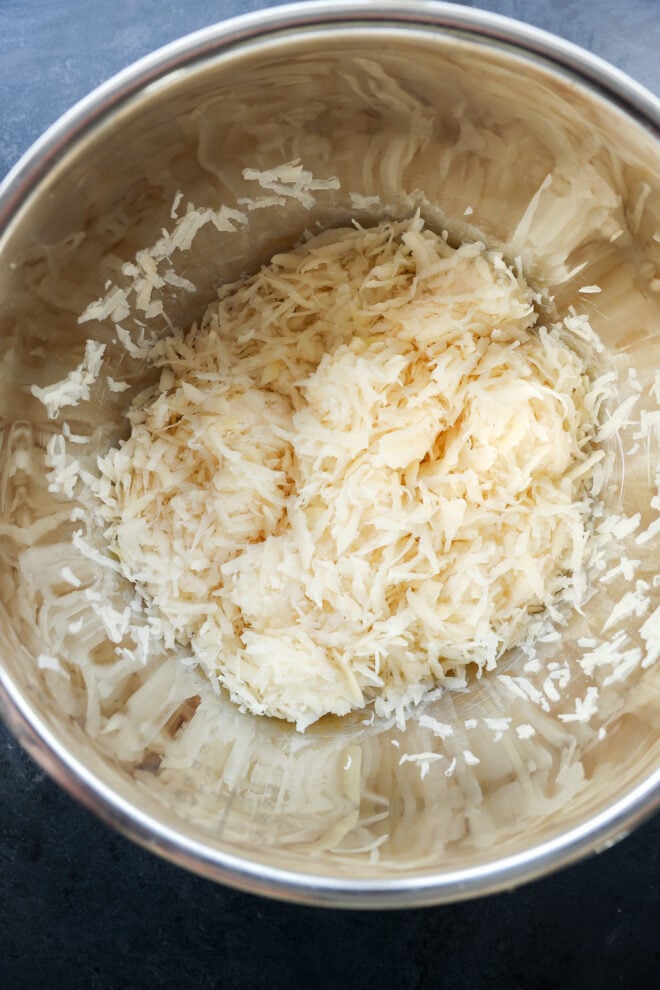 A stainless steel bowl containing freshly grated potatoes, with thin, white shreds spread across the bottom.