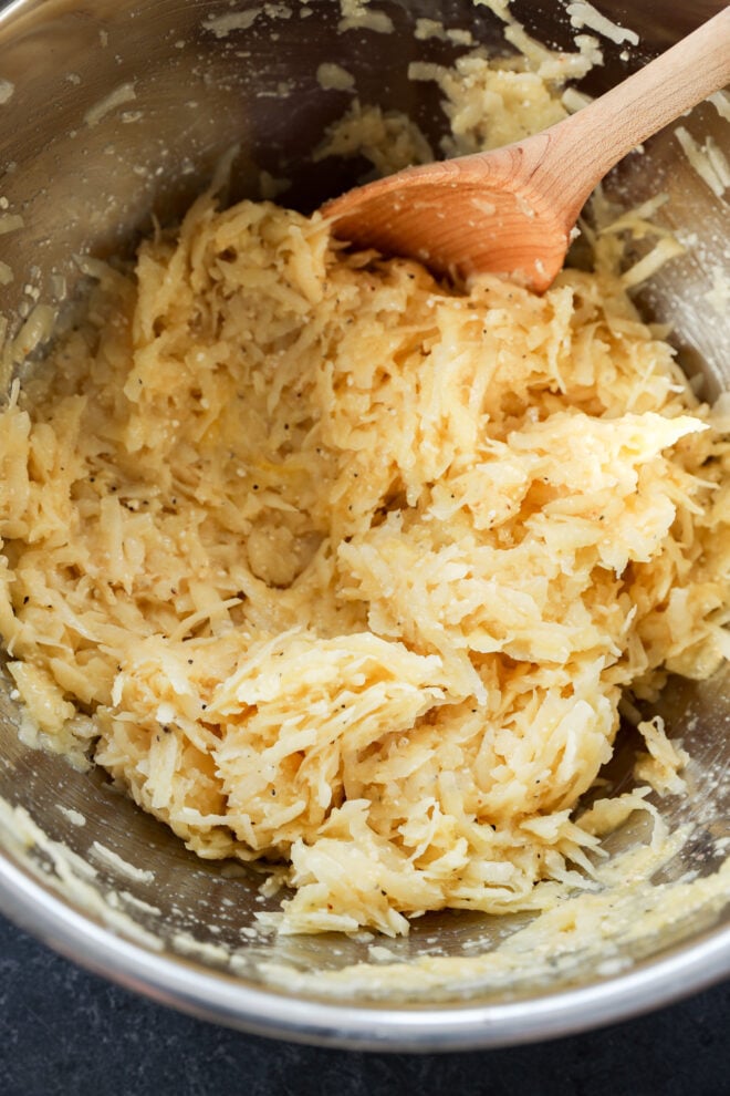 A metal bowl filled with a mixture of grated potatoes and seasonings, being stirred with a wooden spoon. The mixture appears to be prepared for cooking classic Jewish latkes or hash browns.