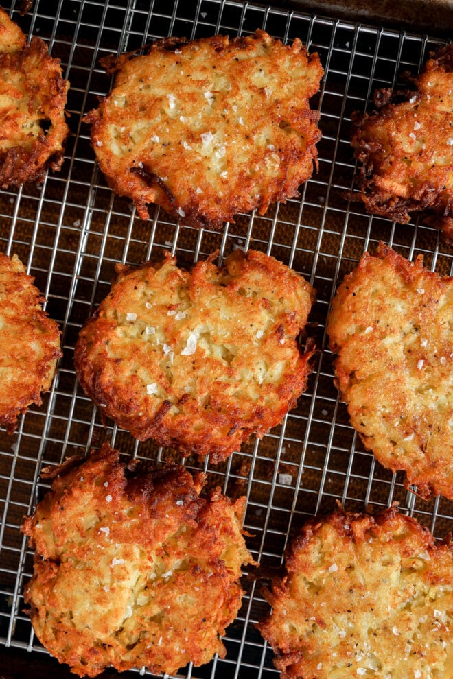 Golden brown Jewish latkes sprinkled with salt are cooling on a wire rack over a dark baking sheet. The crispy texture and uneven edges of the latkes are visible.