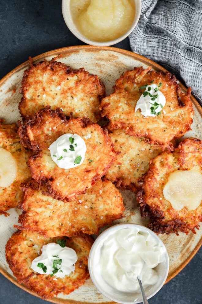 A plate of crispy Jewish latkes topped with sour cream, chives, and applesauce, served with extra sour cream and applesauce on the side. A gray napkin is visible in the background.