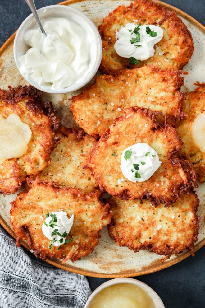 A plate of crispy Jewish latkes topped with sour cream and chopped herbs, served with a bowl of sour cream and a bowl of applesauce on the side. A striped cloth napkin is partially visible in the corner.
