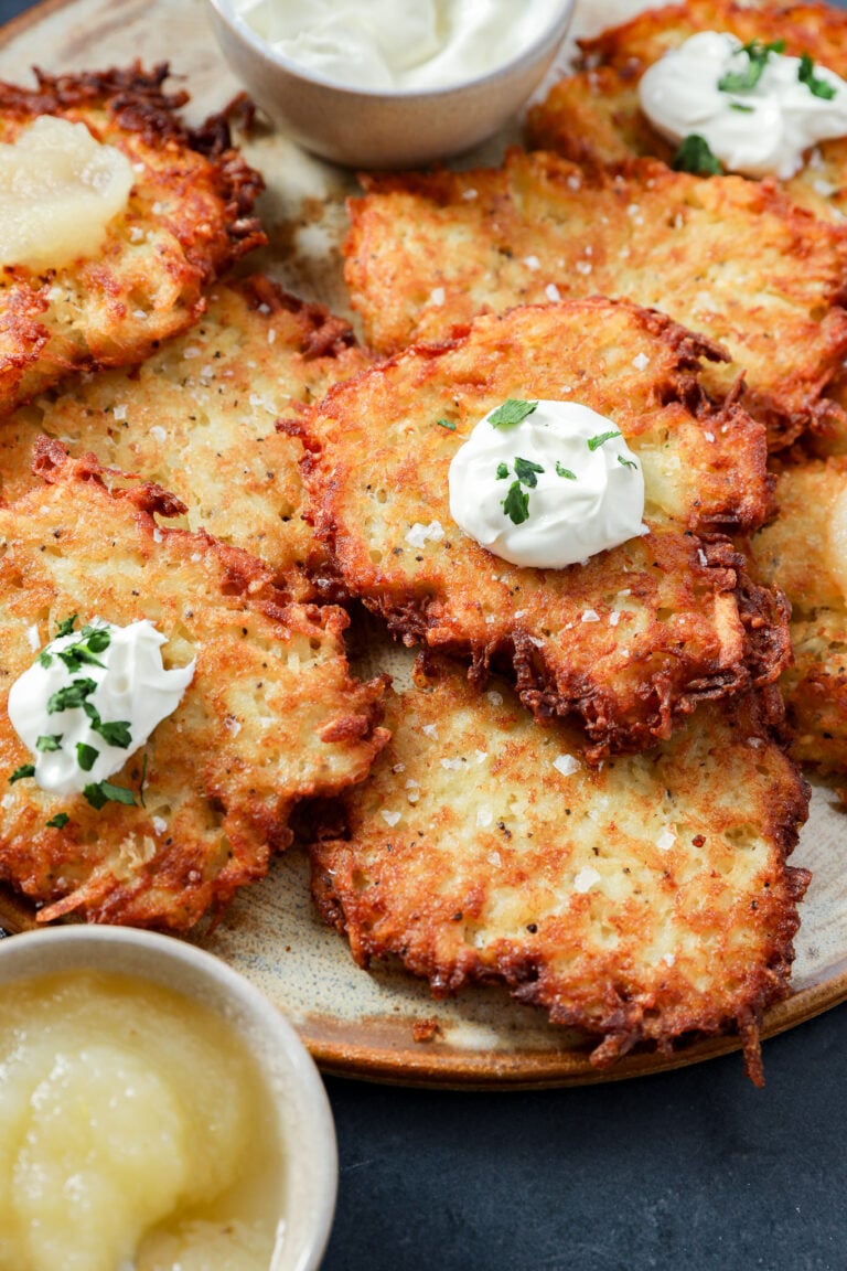 A plate of crispy Jewish latkes topped with dollops of sour cream and herbs, served with bowls of applesauce and sour cream on the side.