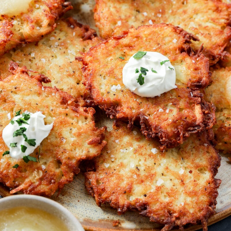 A plate of crispy Jewish latkes topped with dollops of sour cream and herbs, served with bowls of applesauce and sour cream on the side.
