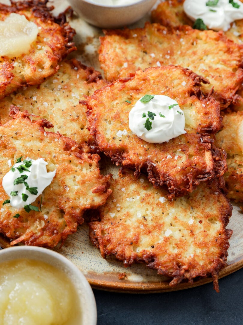 A plate of crispy Jewish latkes topped with dollops of sour cream and herbs, served with bowls of applesauce and sour cream on the side.