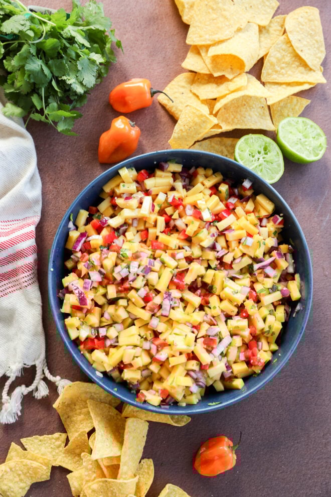 A bowl of fresh mango salsa with diced mango, red onion, and red pepper, surrounded by tortilla chips, halved limes, habanero peppers, cilantro, and a striped cloth on a brown surface.