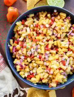 A bowl of mango salsa with diced mango, red onion, red bell pepper, and herbs, surrounded by tortilla chips, a halved lime, habanero peppers, and a white cloth on a brown surface.