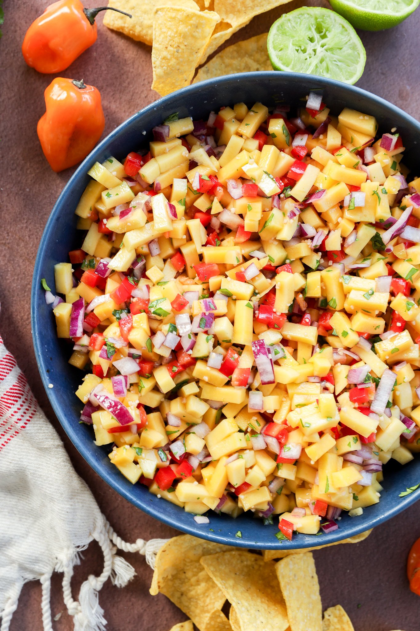 A bowl of mango salsa with diced mango, red onion, red bell pepper, and herbs, surrounded by tortilla chips, a halved lime, habanero peppers, and a white cloth on a brown surface.