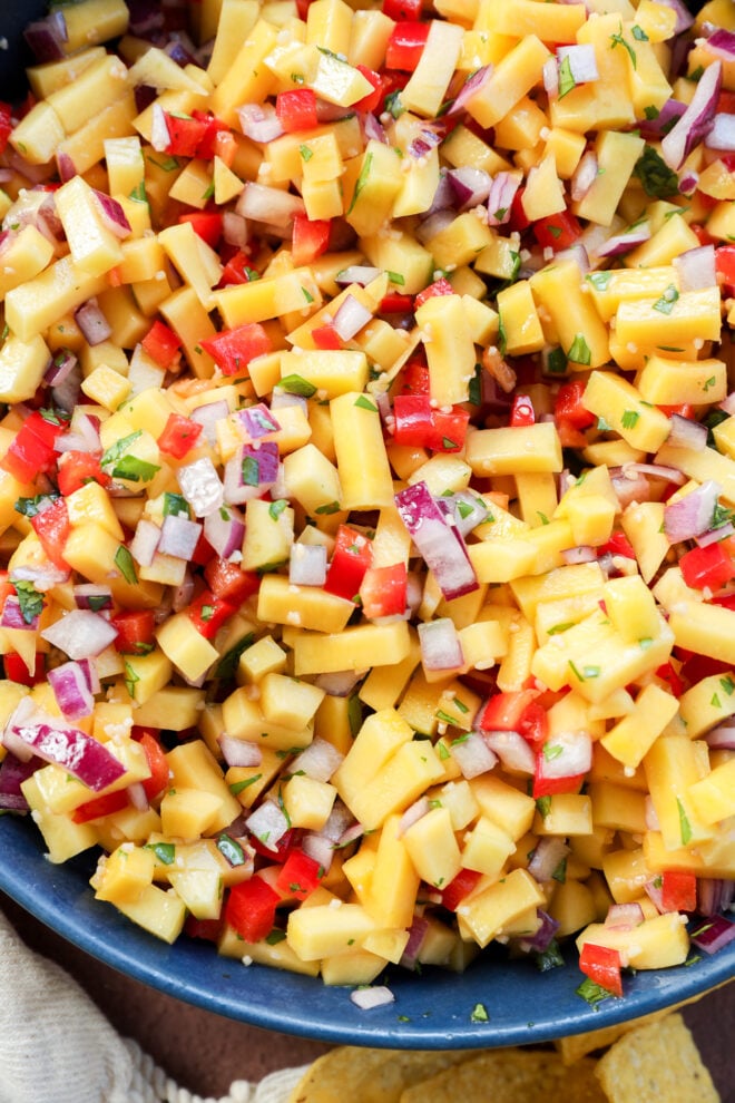 A close-up of a colorful mango salsa with diced mango, red bell pepper, red onion, and cilantro mixed together in a bowl. Tortilla chips are partially visible at the corner of the image.