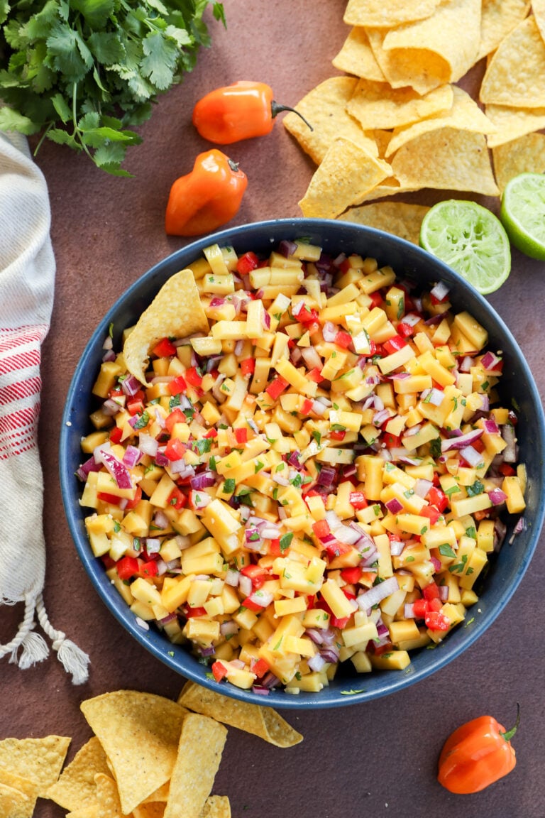 A bowl of mango salsa with diced mango, red onion, red peppers, and herbs, surrounded by tortilla chips, halved lime, fresh cilantro, and whole habanero peppers on a brown surface.