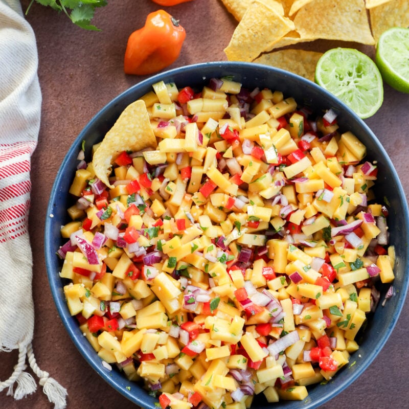 A bowl of mango salsa with diced mango, red onion, red peppers, and herbs, surrounded by tortilla chips, halved lime, fresh cilantro, and whole habanero peppers on a brown surface.