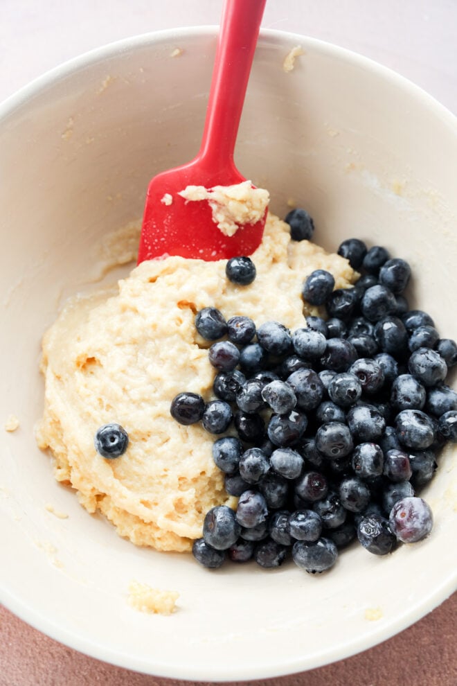 A mixing bowl with mini blueberry muffins batter and fresh blueberries, being stirred with a red spatula.