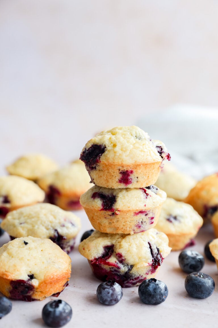 A stack of three mini blueberry muffins is surrounded by more muffins and fresh blueberries on a light surface, with a soft, blurred background.