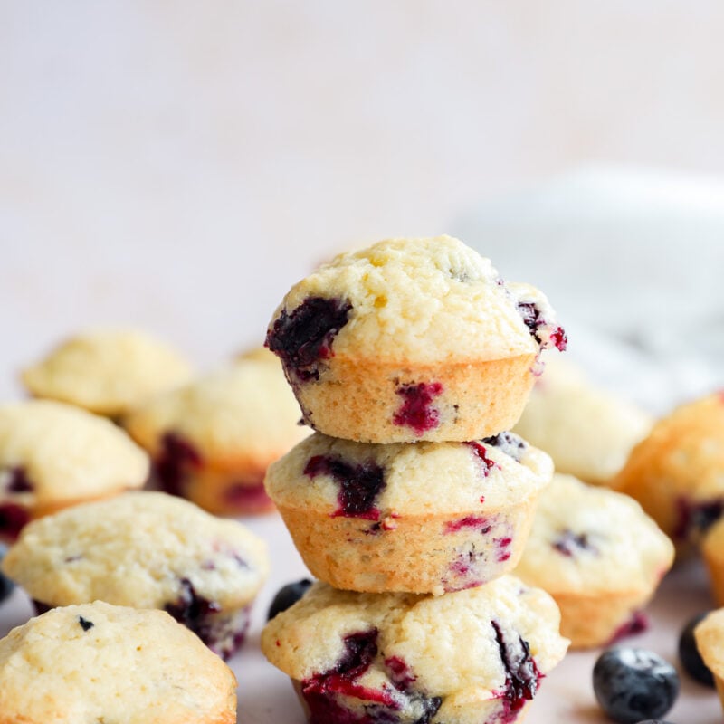 A stack of three mini blueberry muffins is surrounded by more muffins and fresh blueberries on a light surface, with a soft, blurred background.