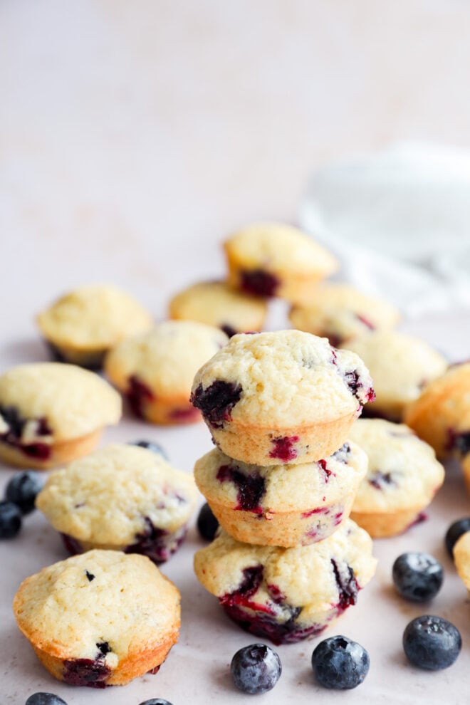 A stack of three mini blueberry muffins is surrounded by more muffins and fresh blueberries on a light surface, with a soft white cloth blurred in the background.