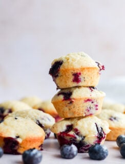A stack of three Mini Blueberry Muffins is in focus, with more muffins and scattered fresh blueberries around them on a light surface. The background is softly blurred.