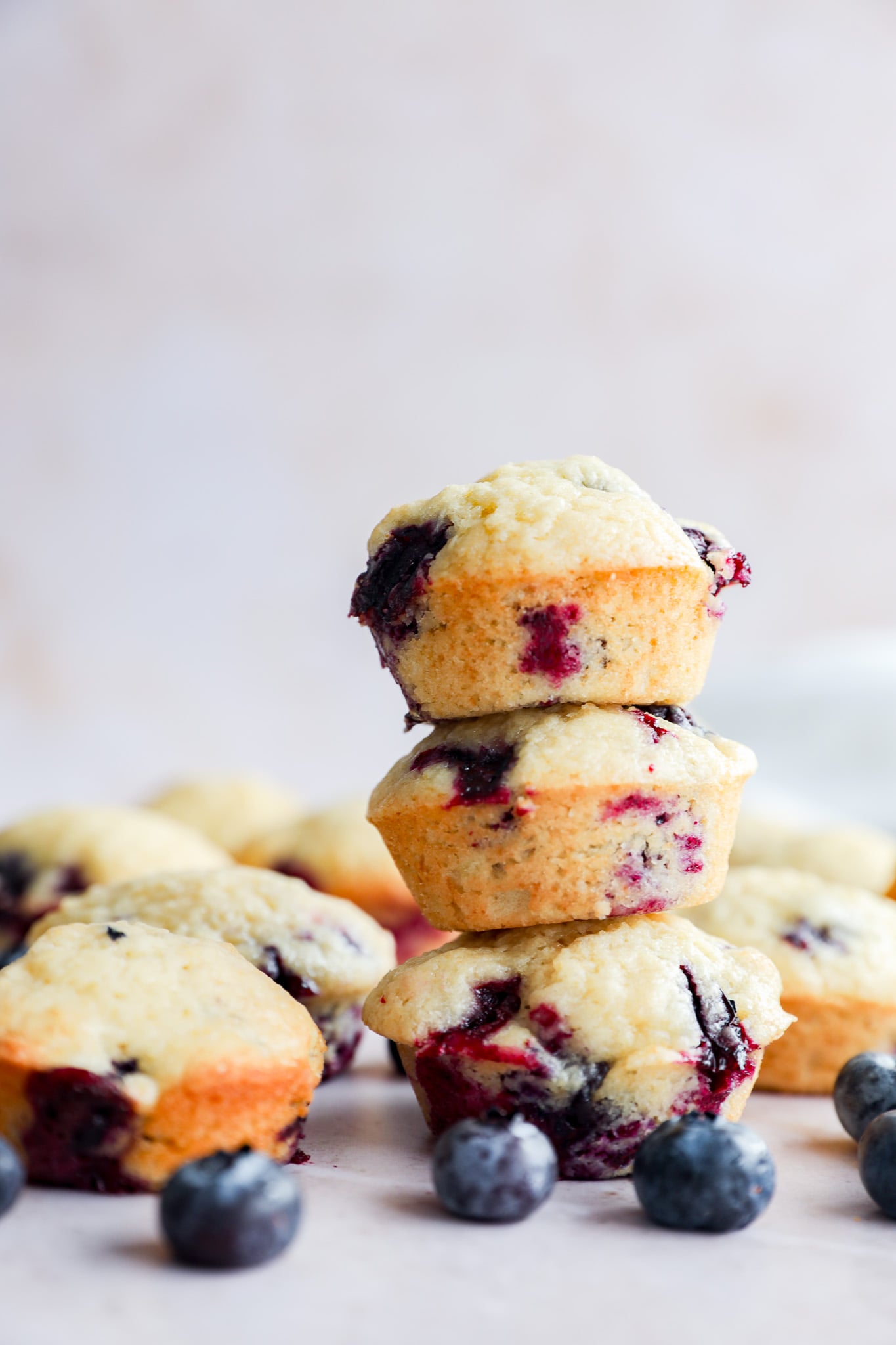 A stack of three Mini Blueberry Muffins is in focus, with more muffins and scattered fresh blueberries around them on a light surface. The background is softly blurred.