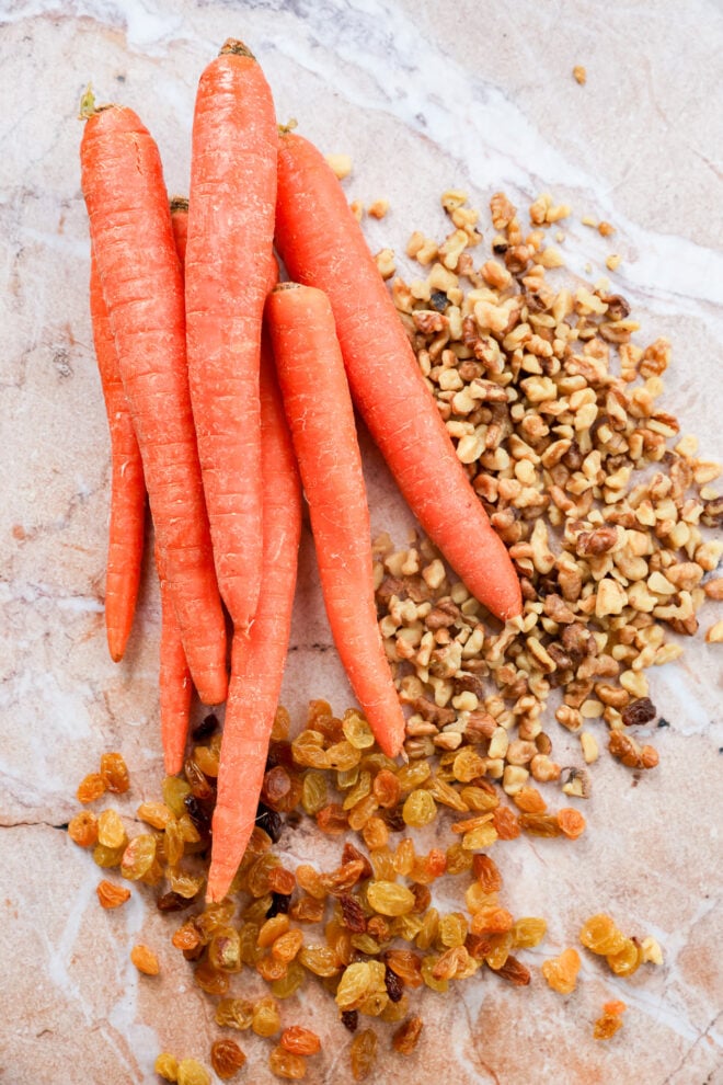 Five whole carrots, a pile of chopped walnuts, and a heap of golden raisins are arranged on a light marble surface with text labels for ingredients. perfect ingredients for a classic carrot cake loaf.