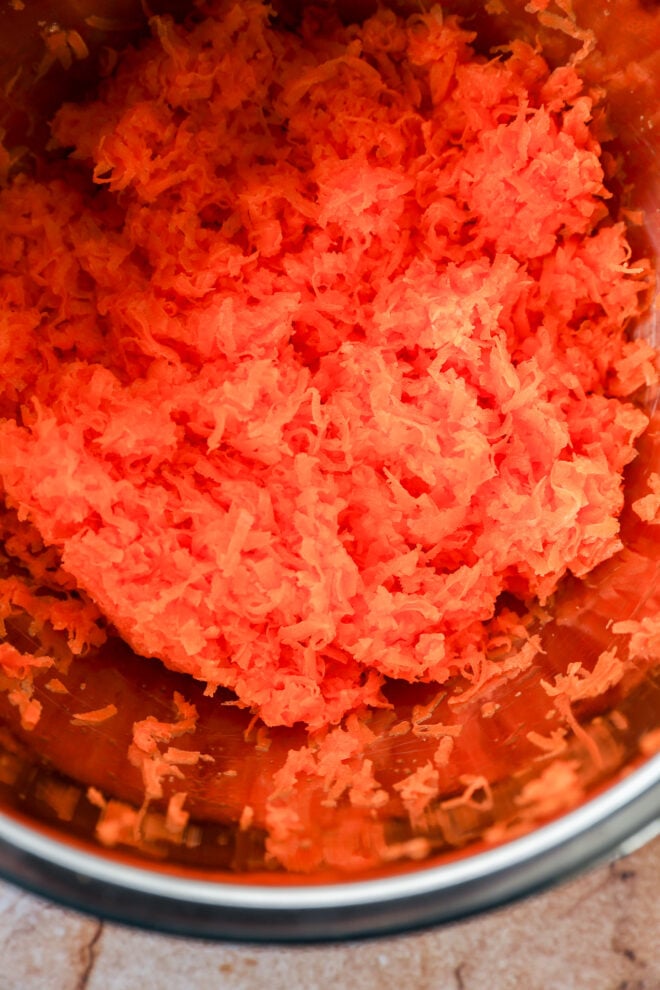 Close-up of finely grated orange carrots in a stainless steel mixing bowl, perfect for preparing a moist carrot cake loaf, sitting on a light-colored countertop.